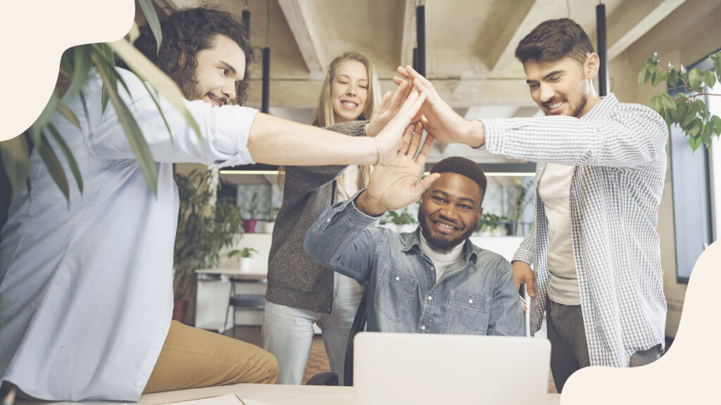 Group of four coworkers in an office giving a high-five around a laptop.