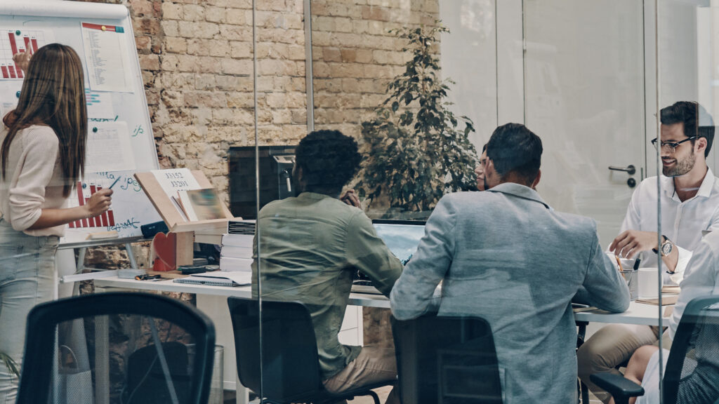 Team in a glass-walled meeting room reviewing charts on a whiteboard and documents on the table.