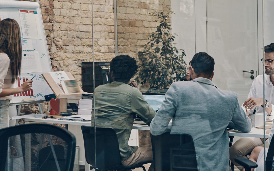 Team in a glass-walled meeting room reviewing charts on a whiteboard and documents on the table.